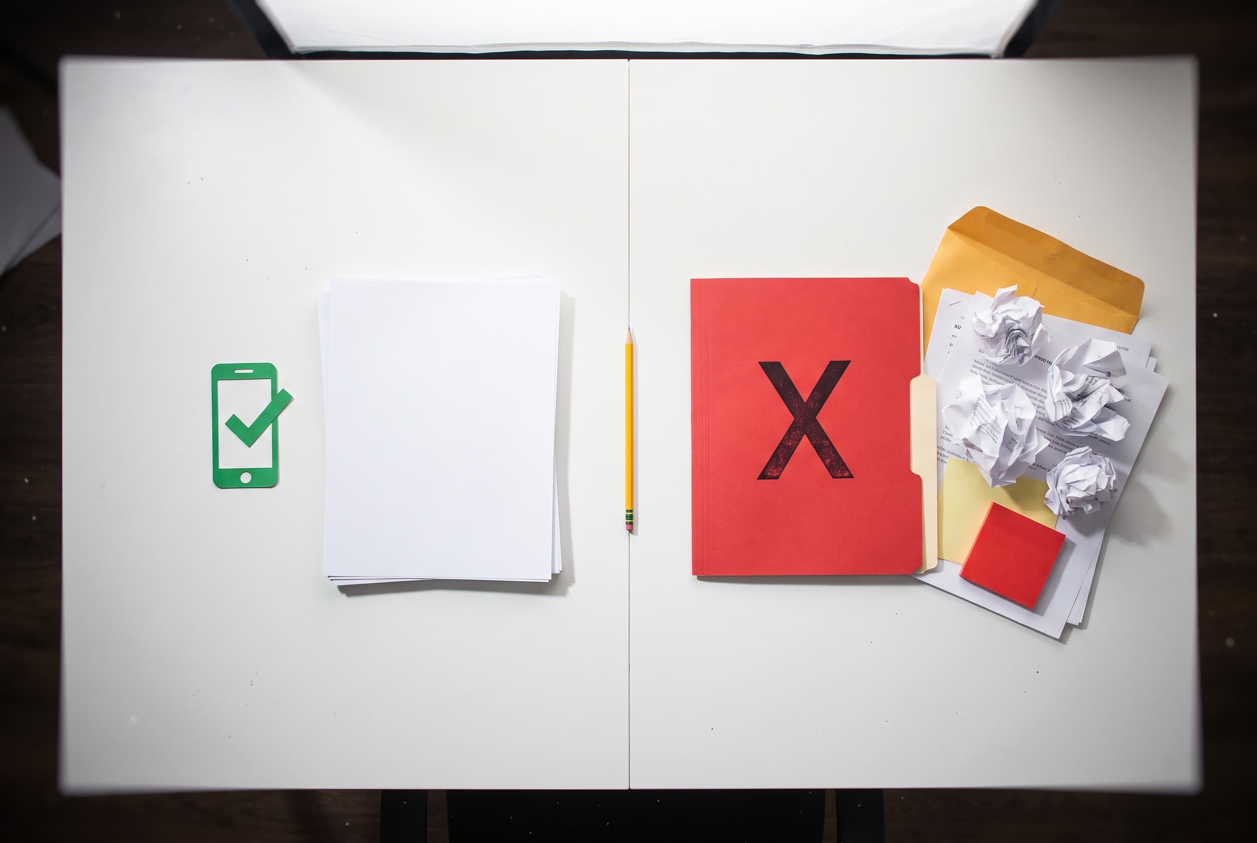 Top-down flat-lay on a clean white desk showing two paths side by side. On the left, a small paper cutout of a smartphone ico